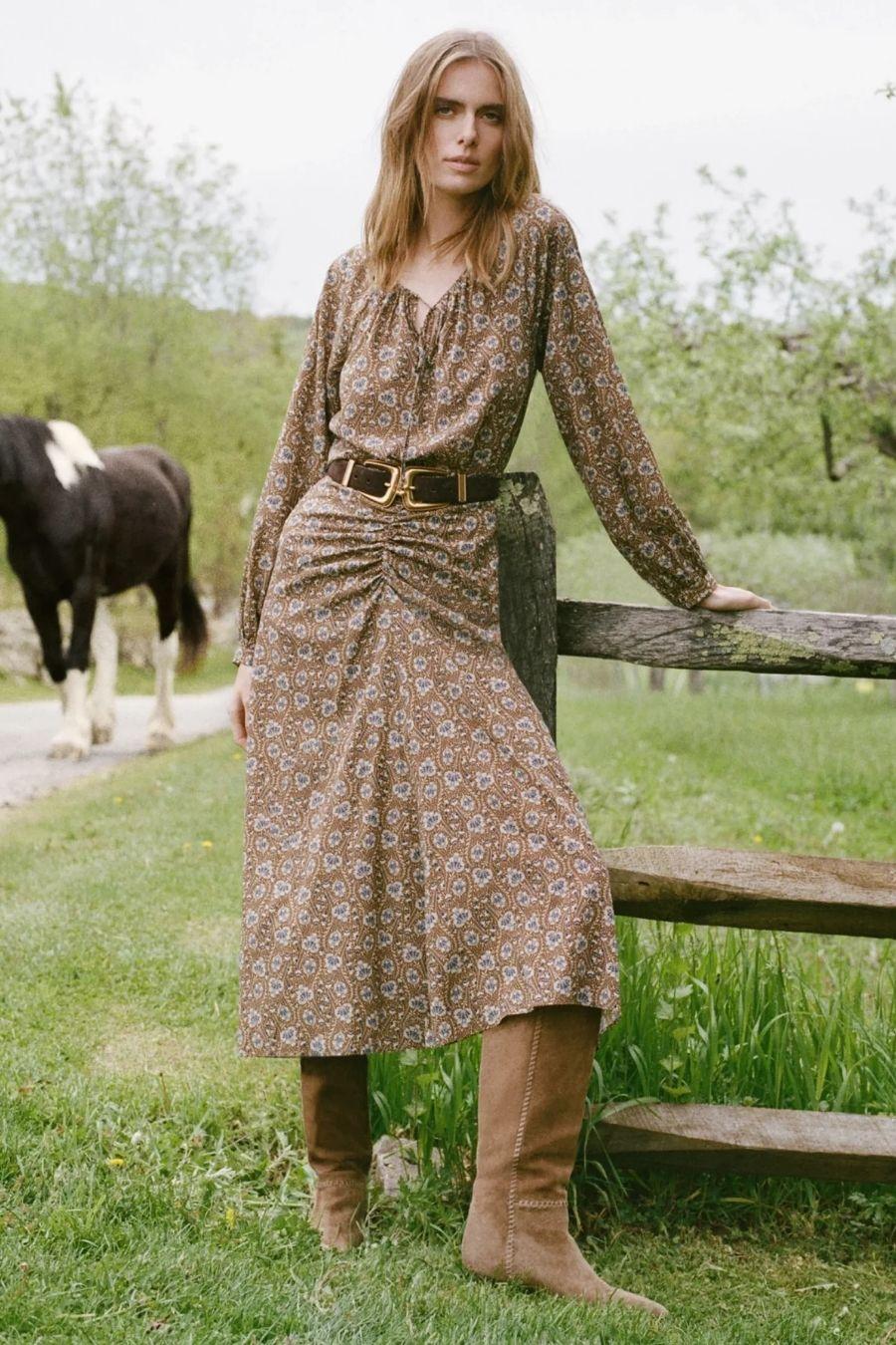Model in floral print midi dress with belt and suede boots, standing by wooden fence with a horse